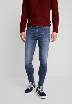 Hombre con jeans ajustados azules, zapatillas deportivas azul marino y blancas, y un suéter texturizado de manga larga color burdeos, de pie frente a un fondo claro y liso.