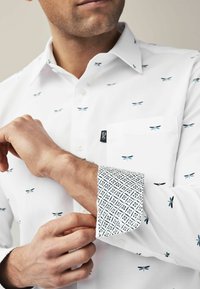 Camisa blanca con estampado de libélulas pequeñas, con un puño estampado en azul marino y blanco. Incluye un bolsillo en el pecho y un diseño de cuello con botones.