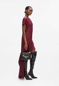 Burgunderfarbenes Kleid mit kurzen Ärmeln und asymmetrischem Saum, kombiniert mit knielangen schwarzen Lederstiefeln und einer schwarzen Handtasche mit silbernen Akzenten.