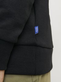 Crna dukserica s rebrastim manžetama i rubom. Ima malu plavu oznaku s natpisom "Jack & Jones" sa strane. Materijal djeluje glatko.
