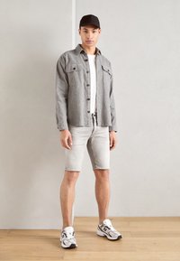 Gray button-up shirt, white t-shirt underneath, light gray denim shorts, and white sneakers with black accents. Man stands against a plain wall.