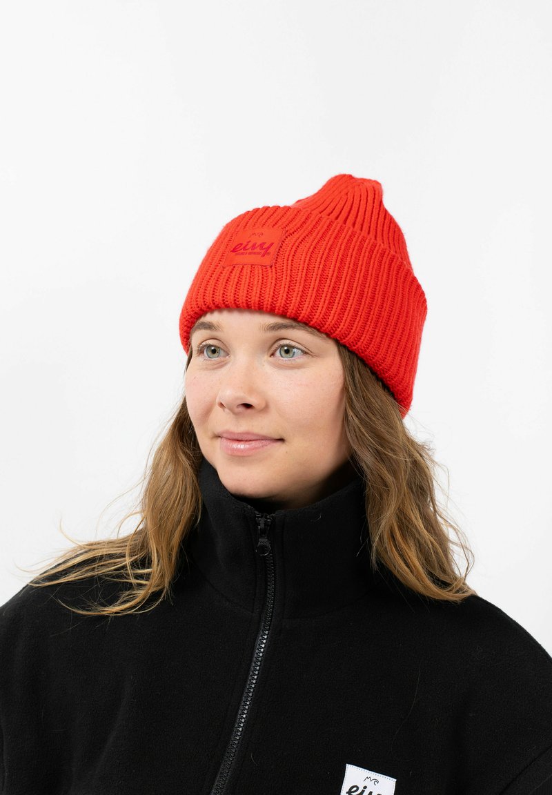 Red knitted beanie with ribbed texture and a small square tag featuring brand name. Worn with a black zip-up fleece jacket.