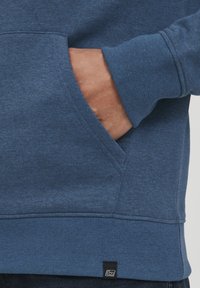 Blå sweatshirt med en forlommes, ribbede manchetter og kant. Materialet har en blød tekstur med et lille mærke på nederste venstre side.