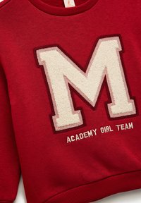 Czerwona bluza z dekoltem ściągaczem. Posiada duże, teksturowane logo "M" w kolorze kremowym i burgundowym oraz wyszywane napisy "ACADEMY GIRL TEAM" poniżej.