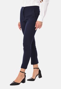 Pantaloni navy su misura con frontale piatto, tasche e vestibilità slim. Abbinati a scarpe con tacco nero e una camicia bianca a maniche lunghe.