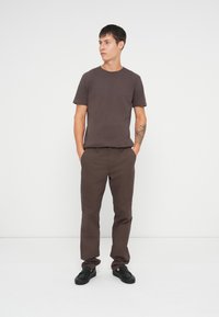 Brun bomulds-t-shirt kombineret med brune bukser og sorte slip-on sko, stående mod en hvid baggrund. Simpel, lige design.