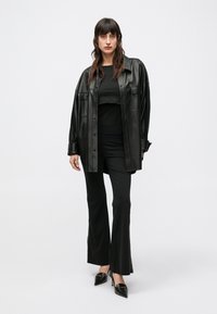 Camicia oversize in pelle nera, top corto nero e pantaloni aderenti neri. Scarpe con tacco alto nere con accenti dorati. Texture lisce e design su misura.