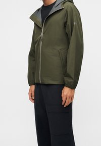 Homme portant une veste à capuche vert olive avec fermeture éclair à l’avant et un pantalon cargo noir, debout devant un fond clair uni.