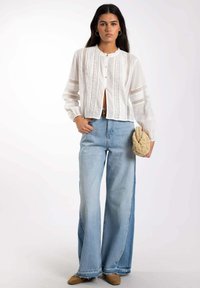 Blusa blanca con detalles de encaje, cierre frontal de botones y mangas largas, combinada con jeans acampanados de tiro alto en un tono azul claro y zapatos beige.