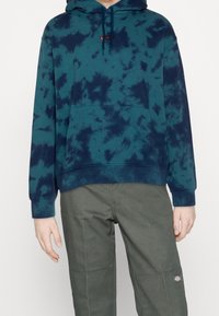 Sweat à capuche tie-dye turquoise en coton, avec une poche avant et une capuche à cordon de serrage. Associé à un pantalon vert olive, mettant en valeur les détails des coutures.