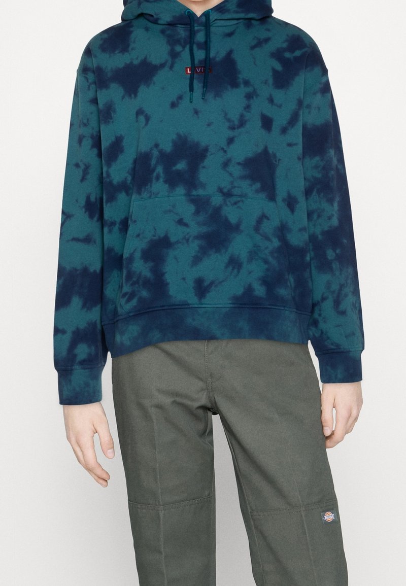 Sweat à capuche tie-dye turquoise en coton, avec une poche avant et une capuche à cordon de serrage. Associé à un pantalon vert olive, mettant en valeur les détails des coutures.