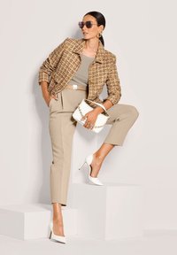 Vrouw in beige broek en tan gekleed vest, met een witte handtas, poseert met één voet op een witte blok tegen een neutrale achtergrond.