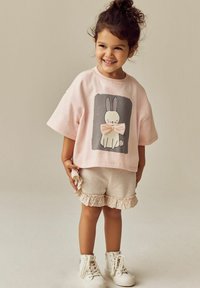 Rosa kortärmad sweatshirt med kanintryck och rosett, ihop med ljusbeige shorts med spetsdetalj och vita högstövelsneakers.