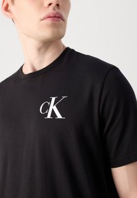 Black cotton t-shirt with a round neckline, featuring a white "cK" logo on the left chest area. Smooth texture and fitted design.