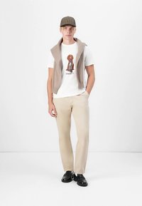 Beežist kootud vest valge graafilise trükiga t-särgi peal, kreemivärvi püksid ja mustad kingad. Mütsil on ruuduline muster.