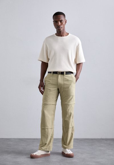 Beige Waffelstrick-Shortsleeve-Oberteil, olivfarbene Cargo-Hosen mit Taschen, schwarzer Gürtel und weiße Sneakers mit braunen Sohlen. Schlichtes, modernes Design.