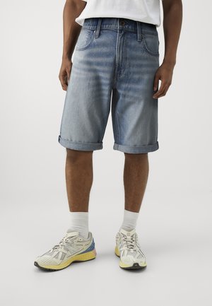 Shorts en denim bleu clair avec ourlet retroussé, présentant un design classique à cinq poches et une légère décoloration. Portés avec des chaussettes blanches et des chaussures de sport.