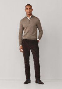 Een bruine ritstrui over een witte kraagblouse, gecombineerd met donkerbruine corduroypantalons en zwarte schoenen, staand tegen een neutrale achtergrond.