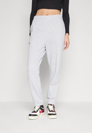 Personne portant un pantalon de survêtement gris clair, un crop top noir à manches longues et des baskets blanches avec des détails noirs et rouges, debout sur fond blanc.