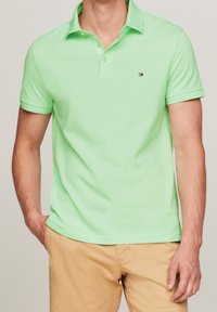 Homme portant un polo à manches courtes vert clair et un pantalon beige, une main dans la poche, sur un fond uni.