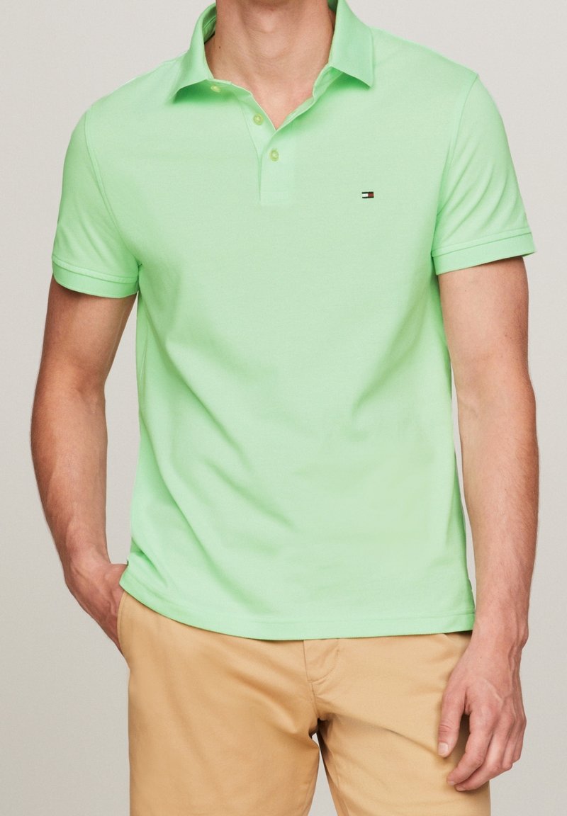 Homme portant un polo à manches courtes vert clair et un pantalon beige, une main dans la poche, sur un fond uni.