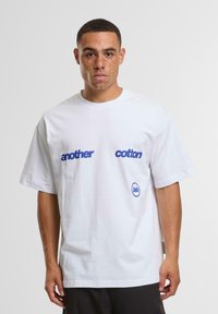 Hvid bomulds T-shirt med blå tekst "another" og "cotton" på fronten, samt et lille blåt logo nederst. Korte ærmer, afslappet pasform.