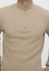 Beżowy dzianinowy sweter z wysokim kołnierzem i półzamkiem z przodu. Całość ozdobiona jest teksturowanym wzorem, posiada dopasowane długie rękawy.