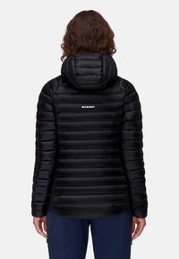 Kvinde iført en sort quilted Mammut hættetrøje, set fra ryggen, stående foran en ensfarvet hvid baggrund.