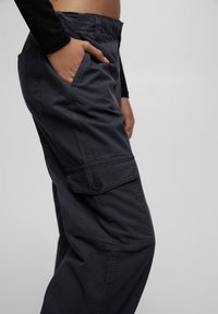 Pantalon cargo gris foncé avec fermeture à boutons, poches latérales et coupe décontractée. Confectionné dans un tissu léger avec des coutures visibles.