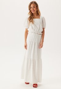 Robe maxi blanche à étages avec des manches bouffantes et un décolleté carré, présentant des motifs à œillets et une taille élastique. Accompagnée de sandales rouges.