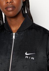 Giacca bomber nera con un motivo in tessuto testurizzato, chiusura a zip, e un logo Nike AIR bianco a contrasto sul petto. Design del colletto a coste.