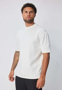 T-shirt blanc à manches courtes avec un col rond haut, présentant un motif subtil de petits points bleus, texture lisse et coupe décontractée.