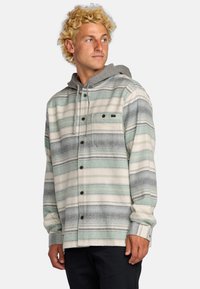 Camisa de hombre con botones que presenta un patrón a rayas horizontales en verde y gris, con una capucha gris y un bolsillo en el pecho. La tela parece suave.