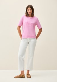 Chemise rose à manches courtes avec "SONO FELICE" imprimé dans un design circulaire, accompagnée d'un pantalon blanc cropped et de sandales marron.
