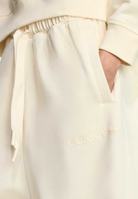 Kremaste Adidas trenirke s elastičnim pojasom, vezicom i bočnim džepovima. Ima utisnuti logo na nozi.