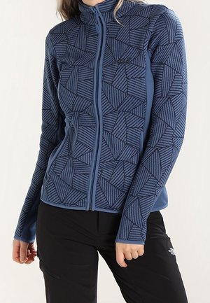Vrouw draagt een blauwe jas met geometrische lijnen en rits en een zwarte broek, staat met één hand bij haar heup.