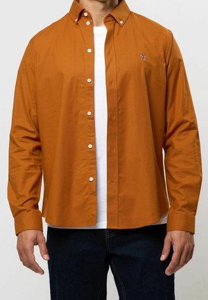 Man die een open burnt orange lang mouwen overhemd draagt over een wit T-shirt en donkere spijkerbroek, staand tegen een effen achtergrond.
