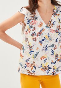 Blomstret bluse med v-hals, korte volangærmer og en cremet baggrund. Har flerfarvede blade og blomster med marineblå, rød og pink detaljer.