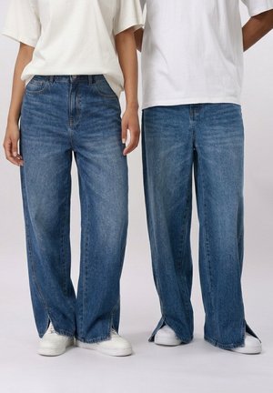 Zwei Personen, die weiße T-Shirts, blaue Jeans mit weitem Bein und vorderen Schlitzen am Saum sowie weiße Turnschuhe tragen, stehen nebeneinander.