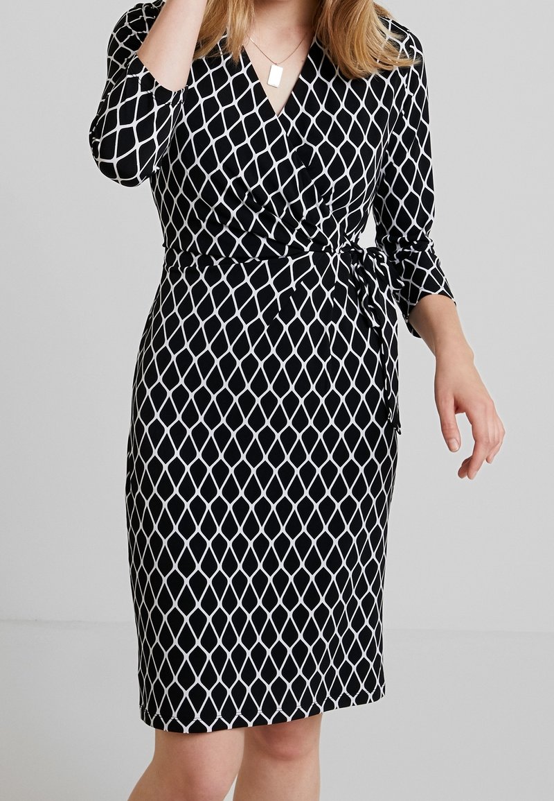 Vestido negro envolvente con patrón de diamantes blancos, mangas tres cuartos, detalle de cintura anudada y falda a la altura de la rodilla. Tejido suave con un ligero brillo.