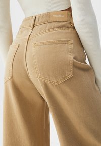 Beige leveled-housut valmistettu denimistä, joissa on korkea vyötärö, kaksi takataskua ja brändilogo vyötäröllä. Rakennekangas.