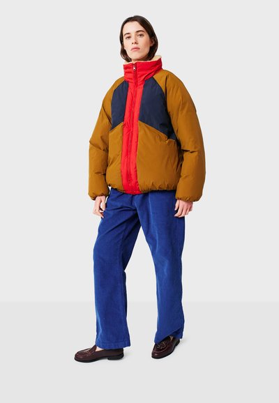 Giacca puffer a blocchi di colore marrone, rosso e blu; colletto alto; texture trapuntata; abbinata a pantaloni di corduroy blu e scarpe scure.