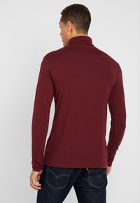 Cămașă lungă bordeaux cu guler înalt, textură netedă și design ajustat, purtată cu blugi negri. Etichetă cu logo vizibilă la tiv.