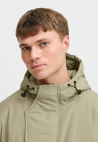 Svetlo khaki bunda s kapucňou má predný zips a šnúrku. Hladká textúra s jemnými švami ponúka moderný dizajn.