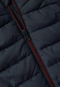 Marinblå kviltad puffjacka med en röd dragkedja och slät, glansig yta. Anmärkningsvärd diamantformad sömnadsdesign över hela jackan.