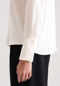 White silk shirt with a buttoned cuff and pleated sleeve detailing, worn over a black bottom, highlighting texture and soft drape.