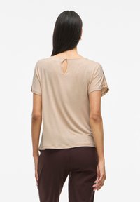 T-shirt beige à manches courtes avec un col rond et un détail cléhole au dos. Tissu doux avec une texture lisse, associé à un pantalon marron foncé.
