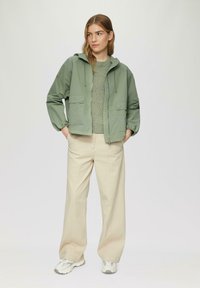 Veste vert clair, de longueur à la taille et avec une capuche, dotée de deux poches avant et de poignets élastiques, portée sur un pull gris texturé et un pantalon beige.