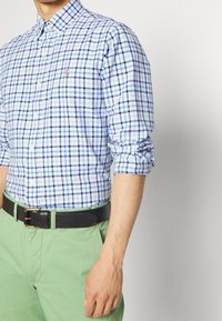 Blå och vit rutig skjorta med knappar, med långa ärmar uppvikt vid mudden, parat med gröna byxor och ett svart bälte.