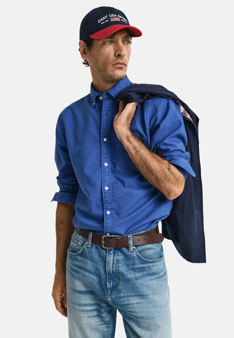 Camicia azzurra a bottoni, jeans azzurri chiari, cintura marrone e cappellino blu navy con visiera rossa, con logo. Giacca appoggiata sulla spalla.